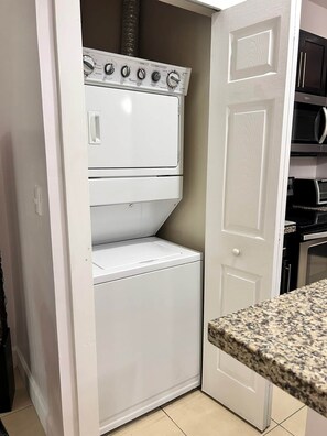 A compact laundry room with a stacked washer and dryer, a door to the right, and a granite countertop. - Sunset 6 Vacations Ground Floor (Coco)