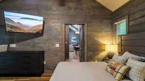 A bedroom with a large bed, a TV mounted on the wall, wooden walls, and a view into the dining area. - Theater! Sauna! Jacuzzi! Firepit! EV Charger (Cosby)