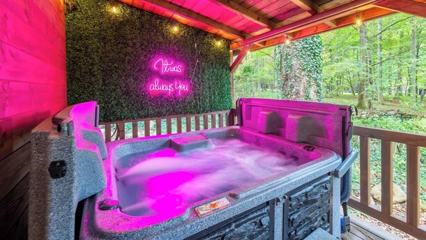 A hot tub with a neon sign, surrounded by a wooden deck and a forested area. - Theater! Sauna! Jacuzzi! Firepit! EV Charger (Cosby)