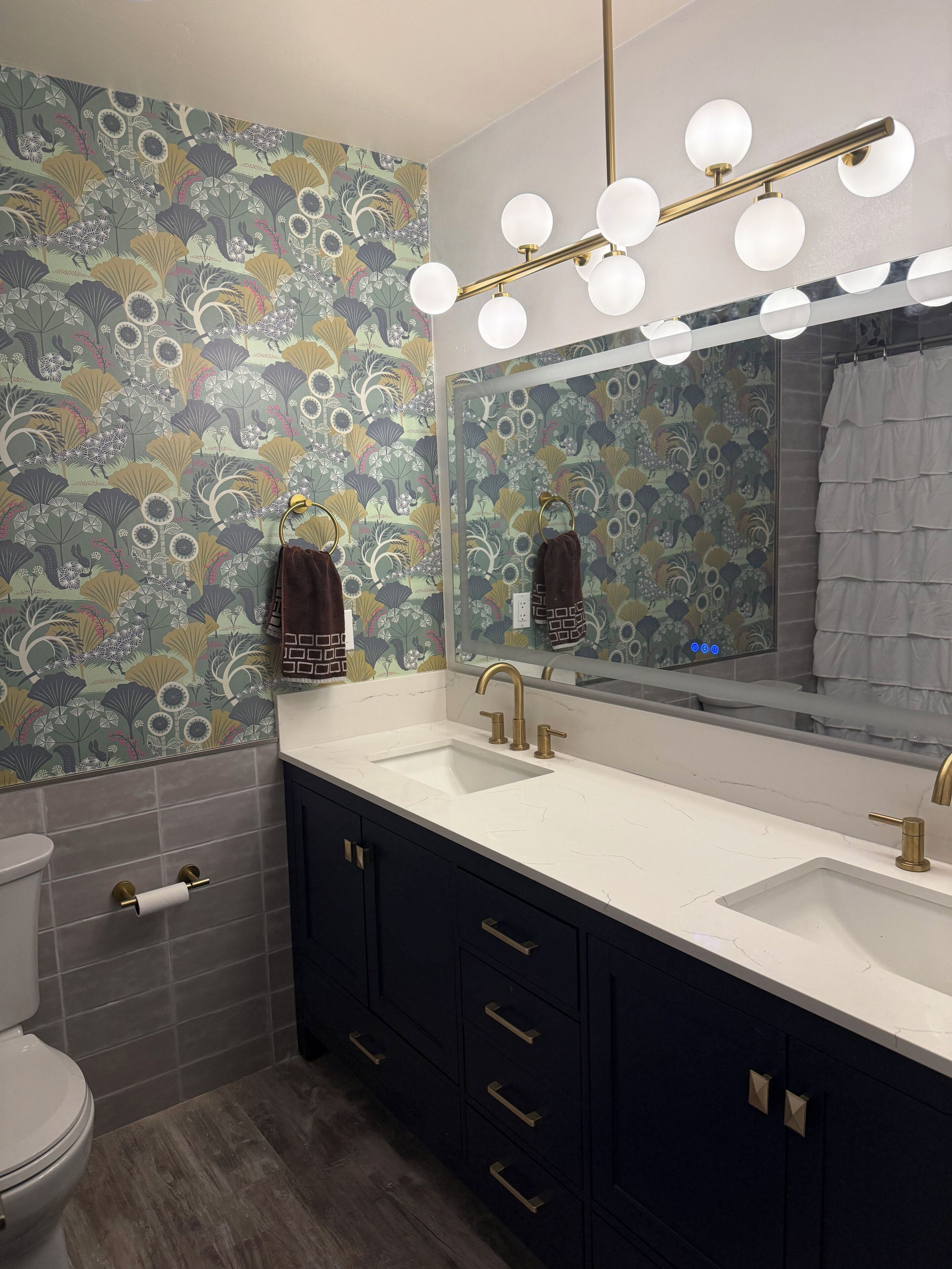 Un bagno con doppio lavandino, uno specchio illuminato, un water e carta da parati decorata.