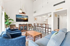 A living room with a large TV, a sailboat decoration, and a dining area with wooden chairs. - Sand Dollar W-210 (Pensacola Beach)