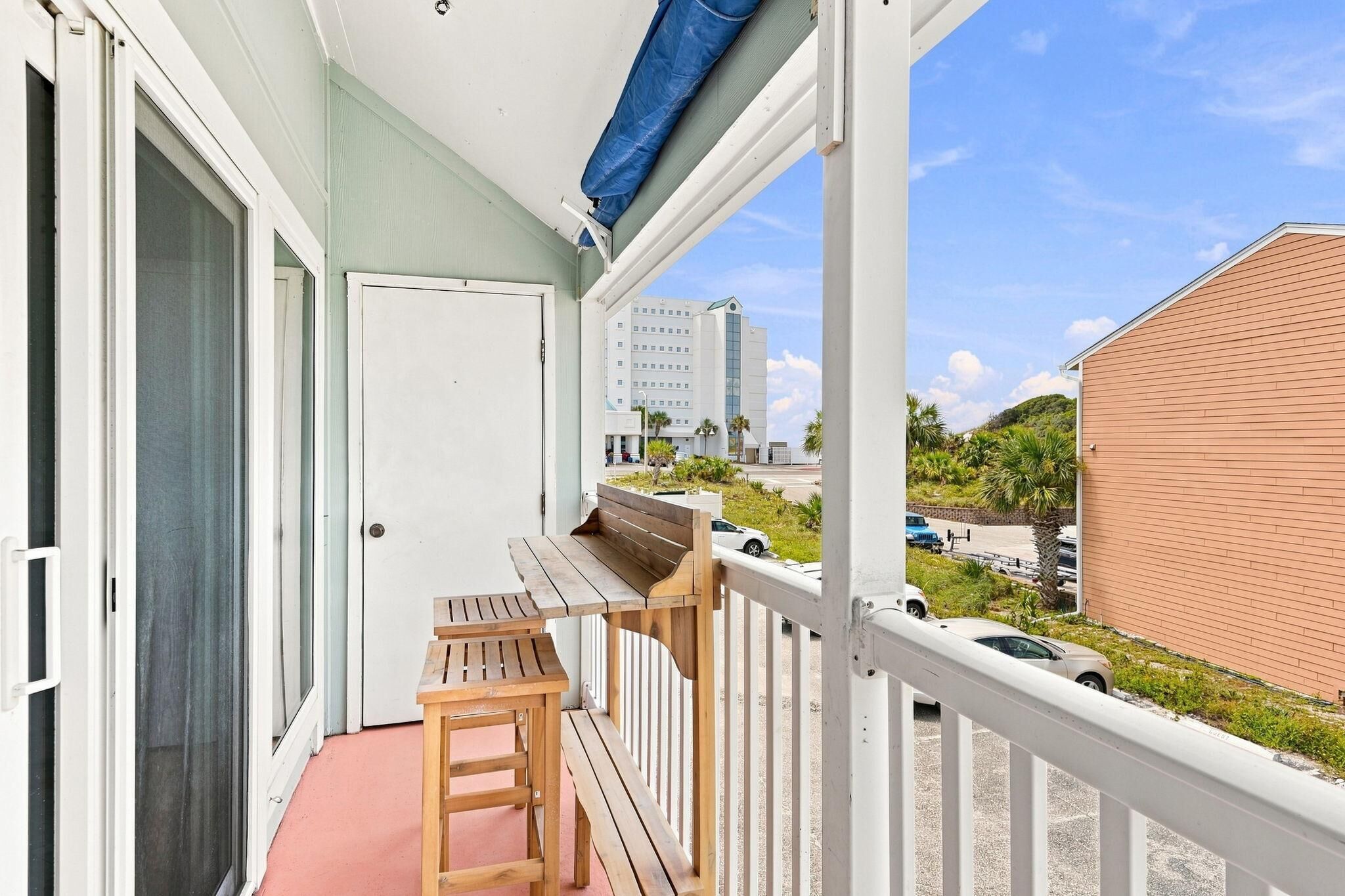 A balcony with a wooden table and chairs, a white door, and a view of a building and parked cars.