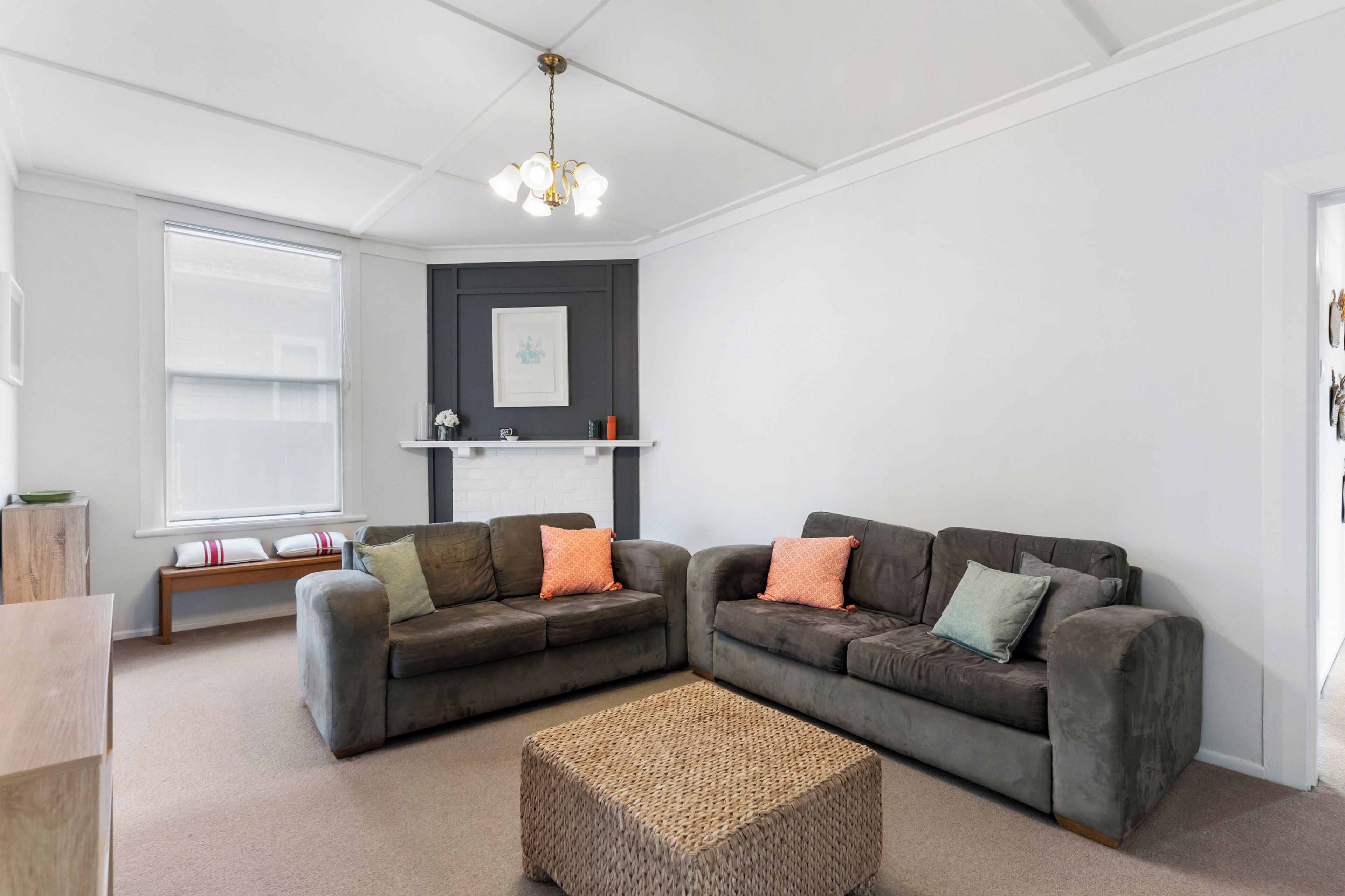 A modern living room with two grey sofas, a woven ottoman, a white ceiling, and a fireplace with a mantel.