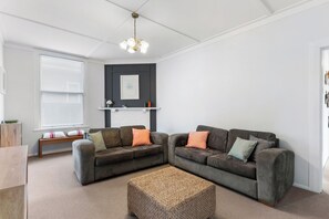 A modern living room with two grey sofas, a woven ottoman, a white ceiling, and a fireplace with a mantel. - Poolside Pause - Auckland Holiday Home (Auckland)
