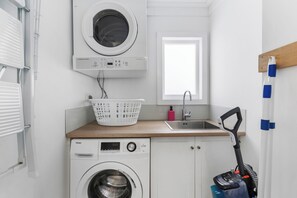 A laundry room with a front-loading washer and dryer, a sink, a window, and a vacuum cleaner. - Poolside Pause - Auckland Holiday Home (Auckland)