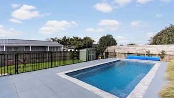 A rectangular swimming pool with a tiled deck, surrounded by a black metal fence, and a wooden deck area in the background.