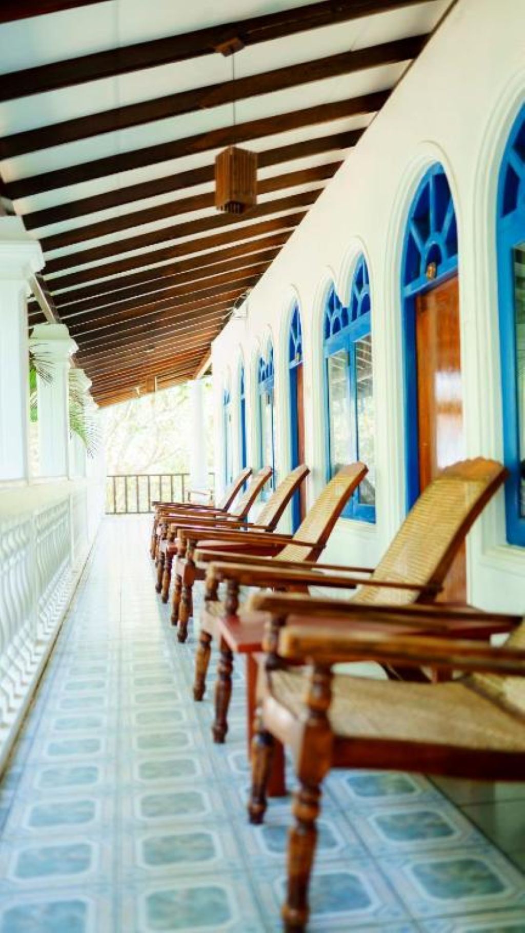 A covered corridor with wooden chairs aligned along the wall, large windows with blue frames, and a patterned floor.