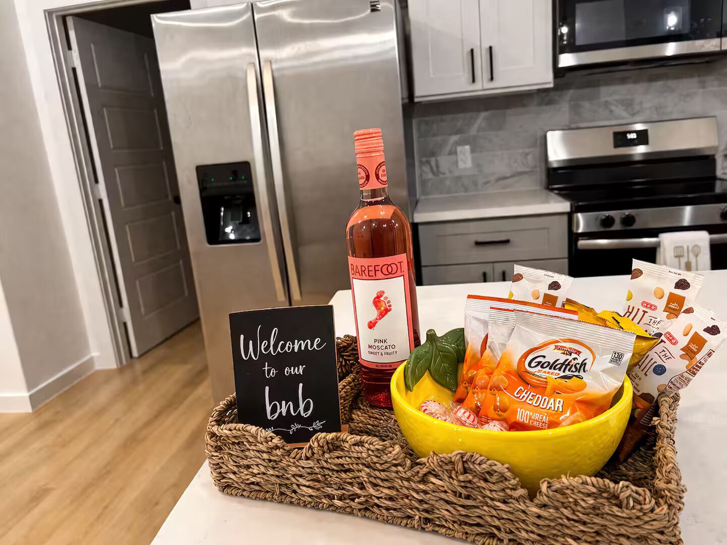 A kitchen with a stainless steel refrigerator, a microwave, and an oven. There is a welcome sign, a bottle of pink Moscato, and a basket with snacks and cheese.
