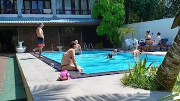 Zona de piscina con un edificio al fondo, una palmera y gente relajándose.