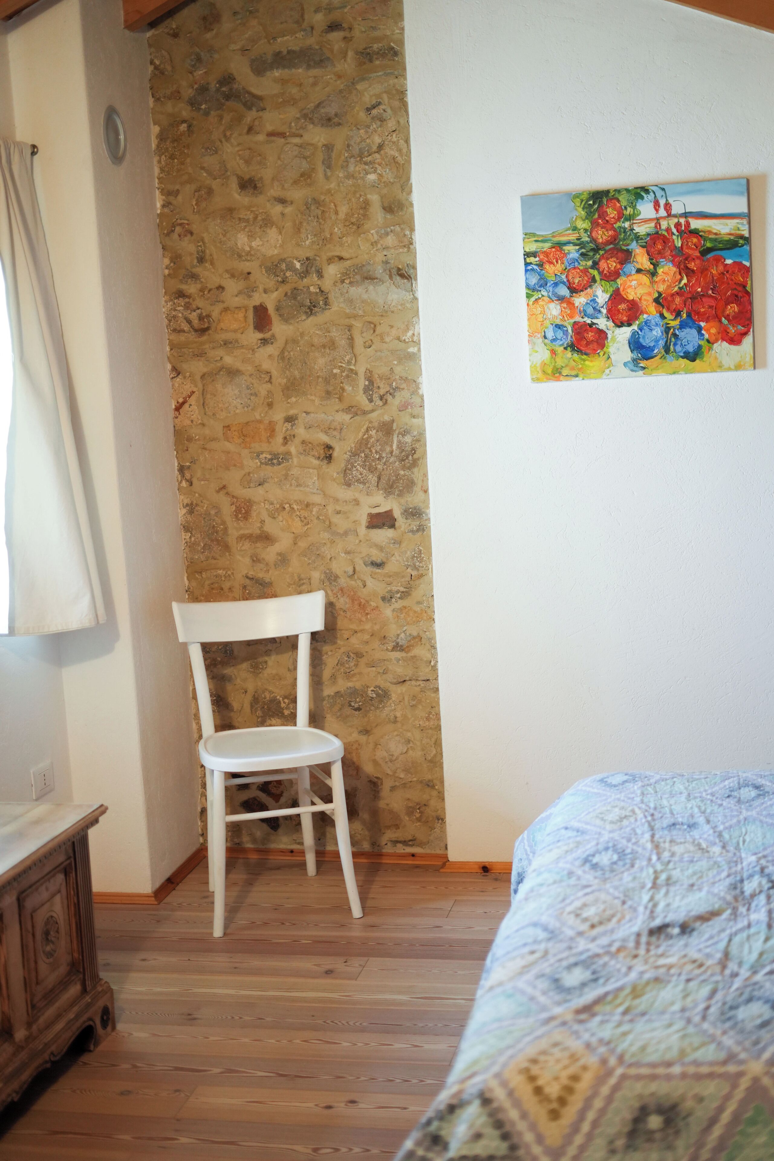 Une pièce avec un mur en pierre, une chaise blanche, un lit recouvert d’une couvre-lit à motifs et un tableau représentant des fleurs accroché au mur.