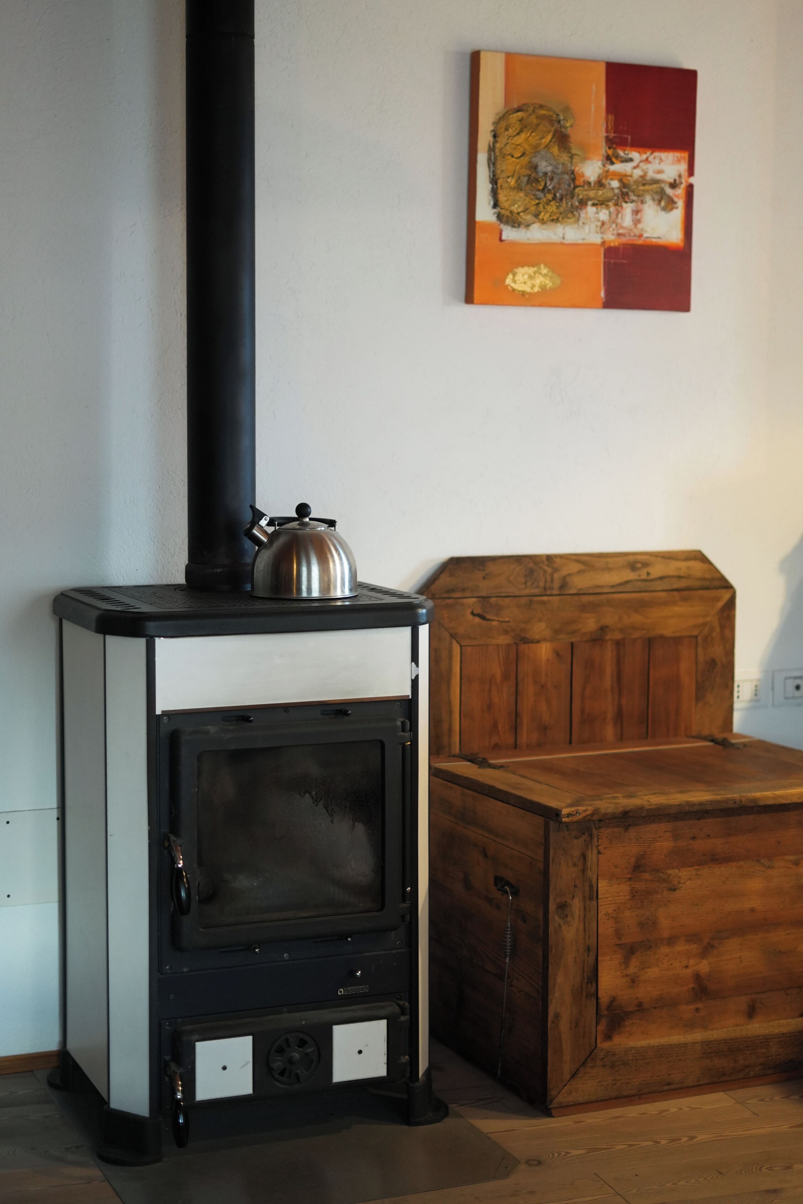 A wood-burning stove with a kettle on top, a wooden chest, and a painting on the wall.