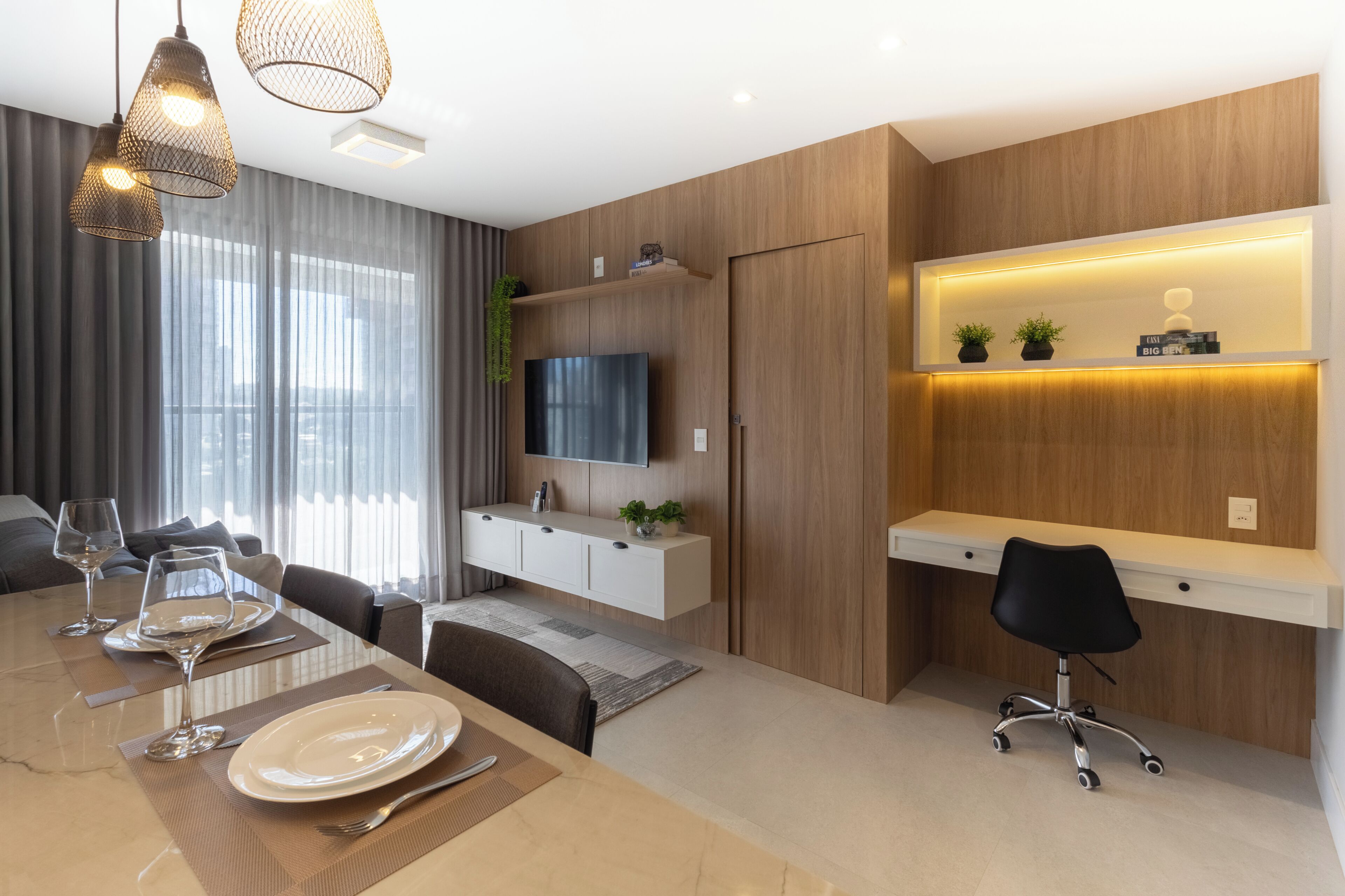 Un espace repas moderne avec une table en bois dressée pour deux personnes, un téléviseur fixé sur un panneau en bois et un bureau avec une chaise.