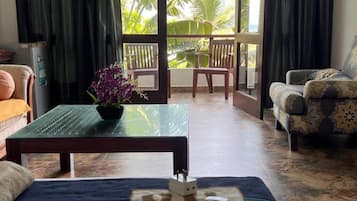 Una habitación con sofá, una mesa de centro con una planta, un control remoto y vistas a palmeras a través de un ventanal.