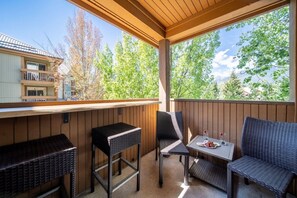A covered outdoor seating area with wicker furniture, a table with a plate of food, and a view of trees and a building in the background. - Whistler Central |Free PKG&Hot Tub,Walk2Everything (Whistler)