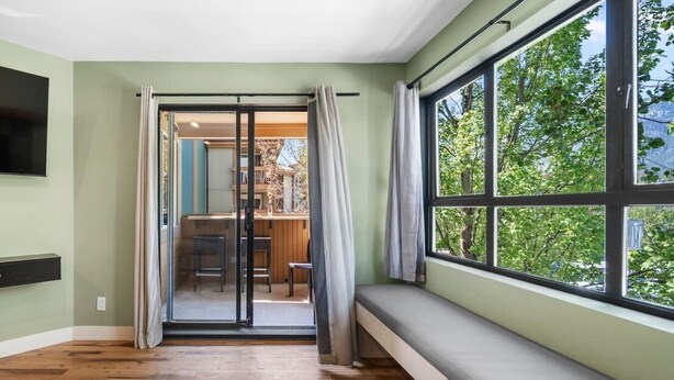 Een kamer met houten vloer, een bank, een schuifdeur naar een balkon en een groot raam met uitzicht op bomen.