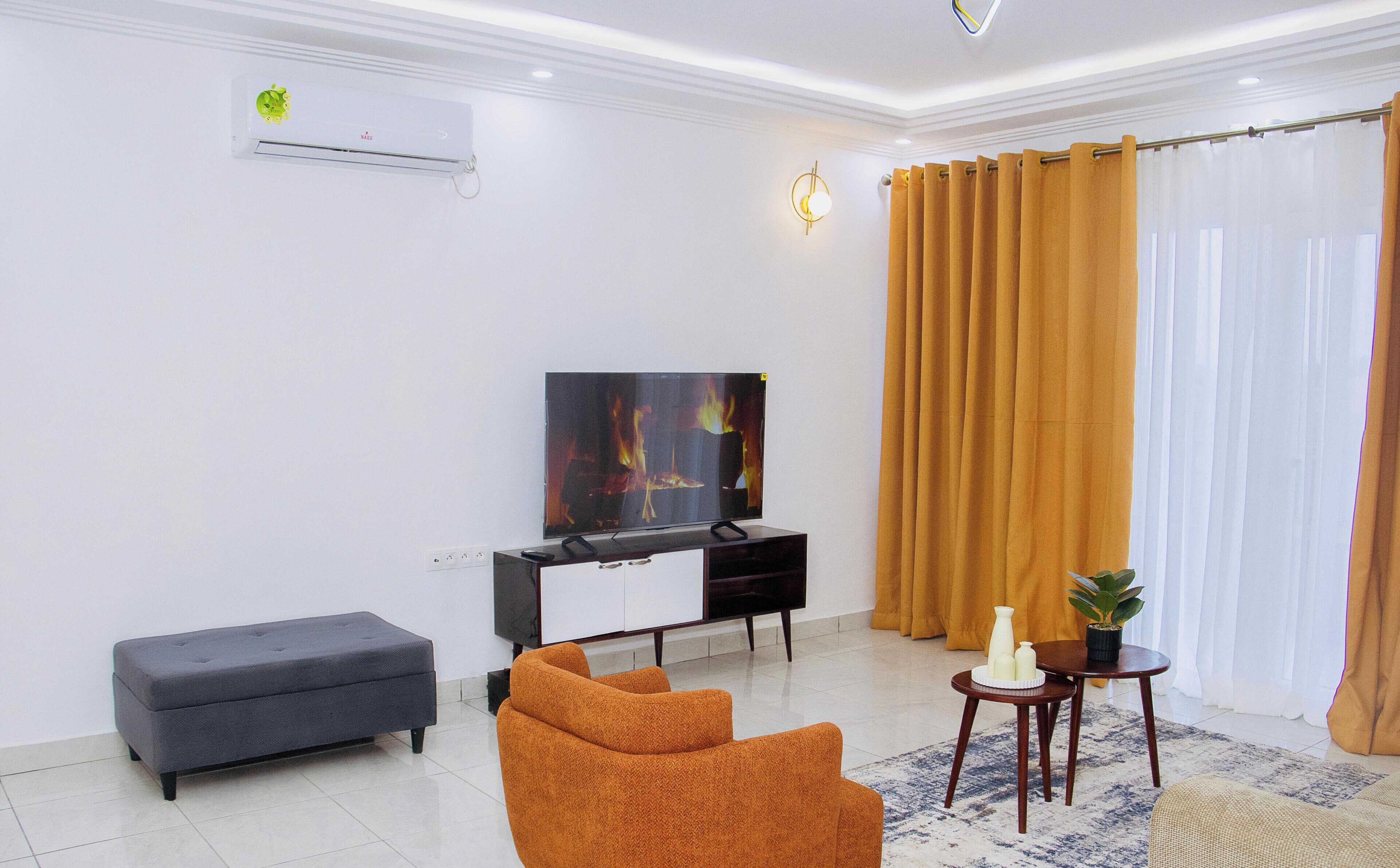 A modern living room with a flat-screen TV, a grey sofa, an orange armchair, a small table with decorative items, and a large window with curtains.