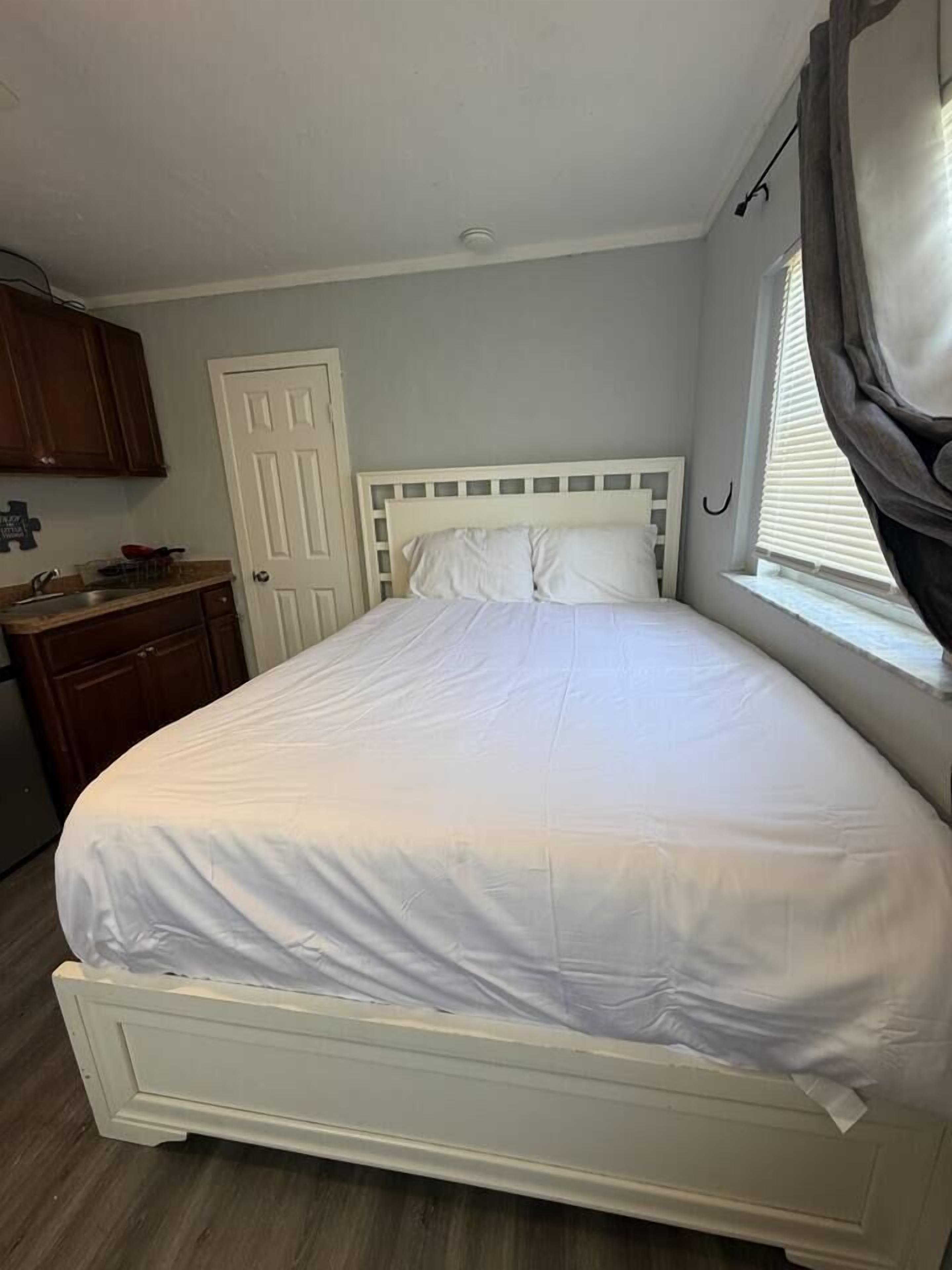 Una camera con un letto singolo, un poggiatesta bianco, una finestra con tende, un comò in legno e una porta.
