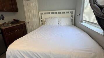 Una camera con un letto singolo, un poggiatesta bianco, una finestra con tende, un comò in legno e una porta.