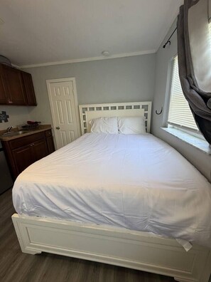 Ein Zimmer mit einem Einzelbett, einem weißen Kopfteil, einem Fenster mit Vorhängen, einem Holzschrank und einer Tür.