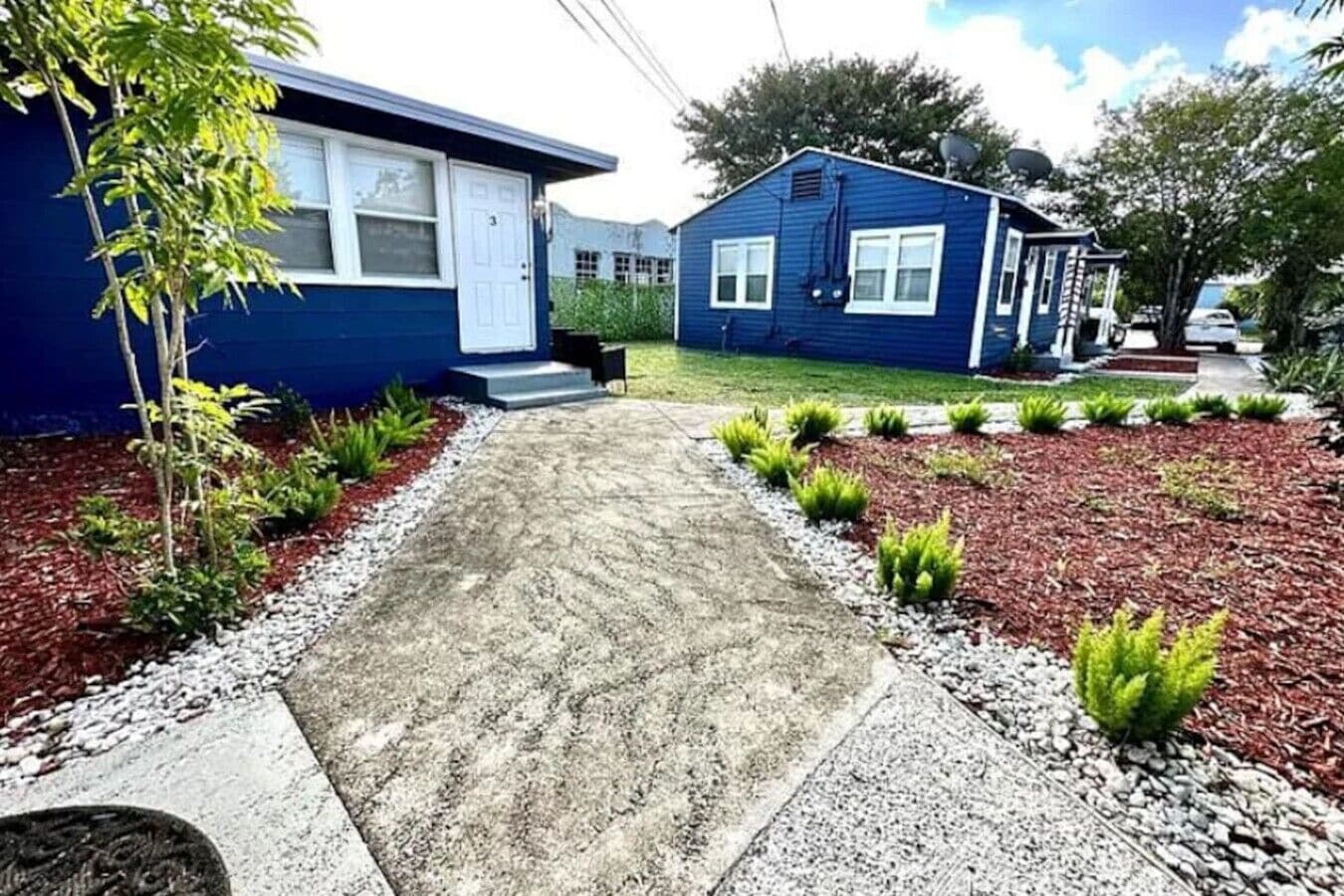 Un immobile residenziale con vialetto in cemento, un piccolo giardino anteriore con pacciamatura rossa e piante verdi, e una casa blu con una porta bianca.