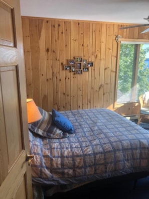 Ein Schlafzimmer mit Holzwand, einem Bett mit Karomuster-Überwurf, einem blauen Kopfkissen und einer kleinen Foto-Collage an der Wand.