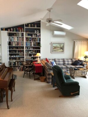 Un salon comprenant une bibliothèque, un ensemble de canapés, un fauteuil vert, un piano et un ventilateur de plafond.