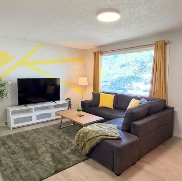 A living room with a flat-screen TV on a white stand, a dark brown sofa with yellow cushions, a small wooden coffee table, and a green rug. - Modern Cozy 2 BR Retreat Near DT & CTrain (Calgary)