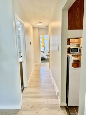 A narrow hallway with a wooden floor, leading to a bedroom with a bed and a nightstand. A microwave is visible in the kitchen area. - Modern Cozy 2 BR Retreat Near DT & CTrain (Calgary)