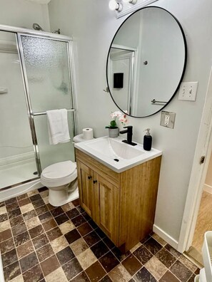 A bathroom with a glass shower enclosure, a white toilet, a wooden vanity with a sink, and a large mirror. - Modern Cozy 2 BR Retreat Near DT & CTrain (Calgary)
