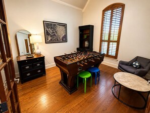 A room with a wooden floor, a foosball table, a chair, a lamp, a map on the wall, and a window with blinds. - 5BR and 3.5 BA in Lower Greenville Home with Game Room Sleeps 14 (Dallas)