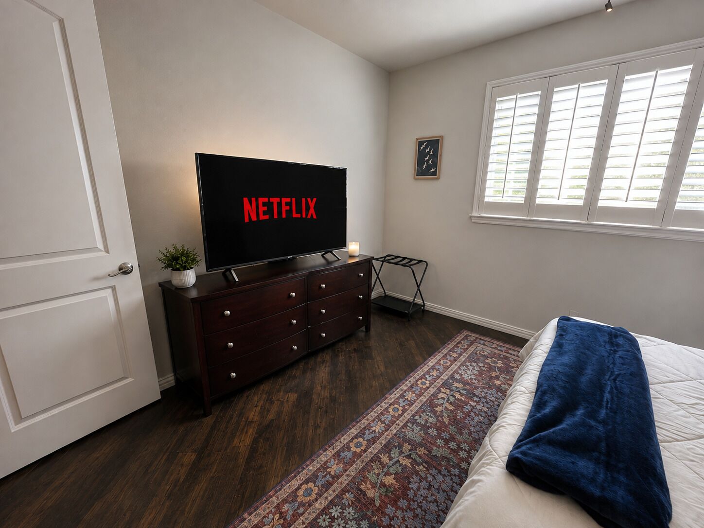 A bedroom with a bed, a dresser, a television displaying 'NETFLIX,' a window with blinds, and a small plant.