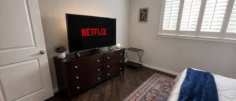 A bedroom with a bed, a dresser, a television displaying 'NETFLIX,' a window with blinds, and a small plant.