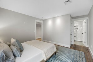 A bedroom with a bed, a bedside table, a mirror, and a door leading to a bathroom with a white tiled shower. - Modern Luxury Commercial St Condo • 3 En-Suite BRs (Provincetown)