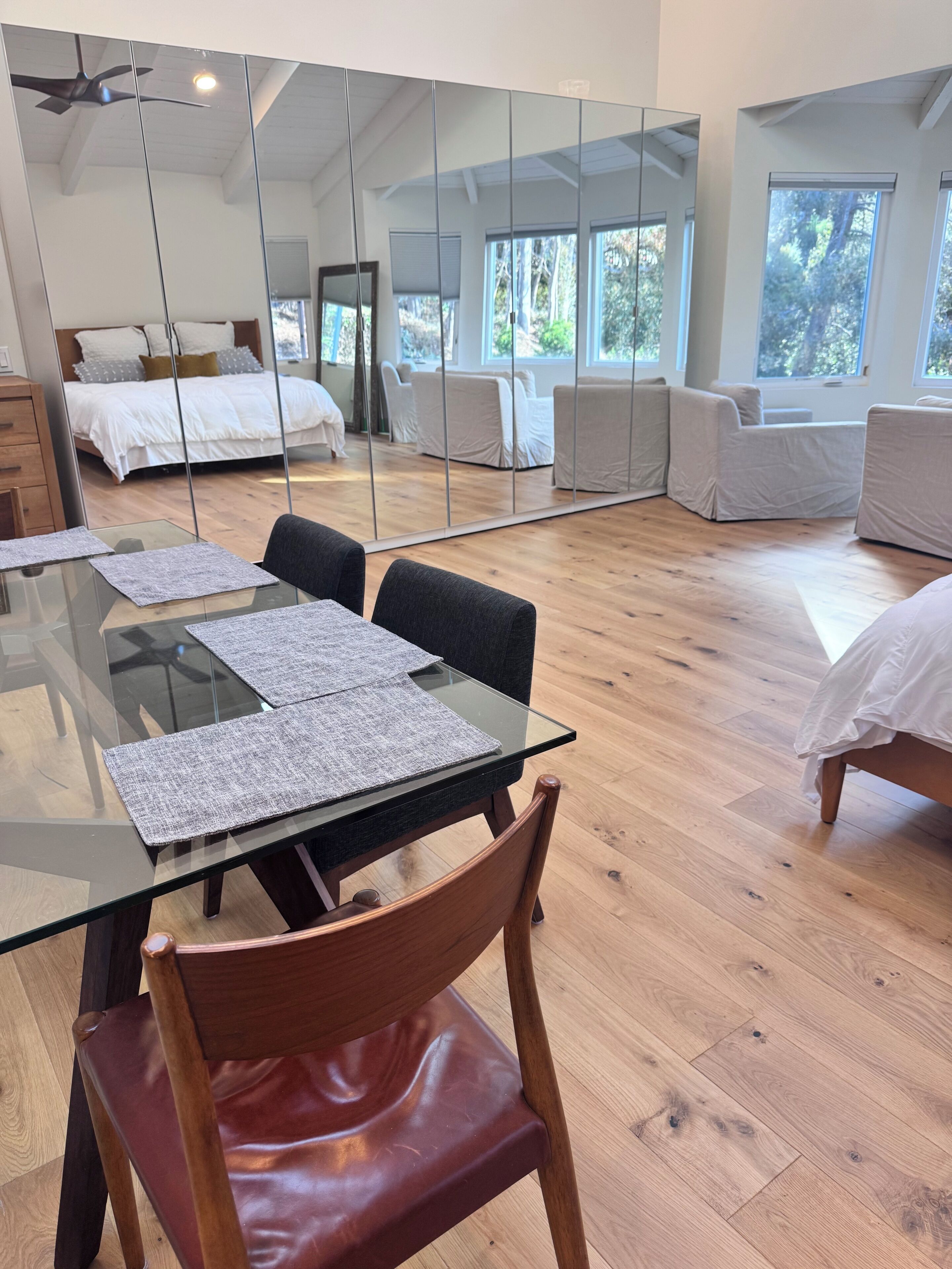Una moderna camera d'albergo con un tavolo da pranzo in vetro, una sedia di legno, un letto e un grande specchio che riflette l'ambiente.