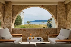 A stone-walled room with a view of a body of water and mountains. There are two sofas and a small table with a glass of orange juice and a smartphone.