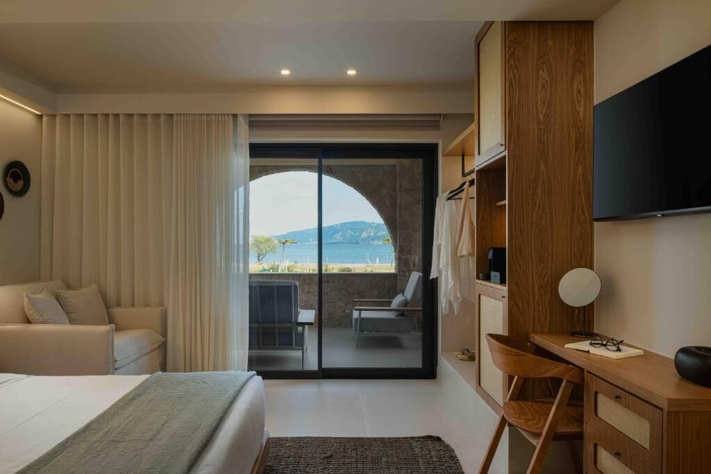 A hotel room with a bed, a desk, a chair, a TV, and a view of the sea through a glass door.