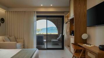 A hotel room with a bed, a desk, a chair, a TV, and a view of the sea through a glass door.