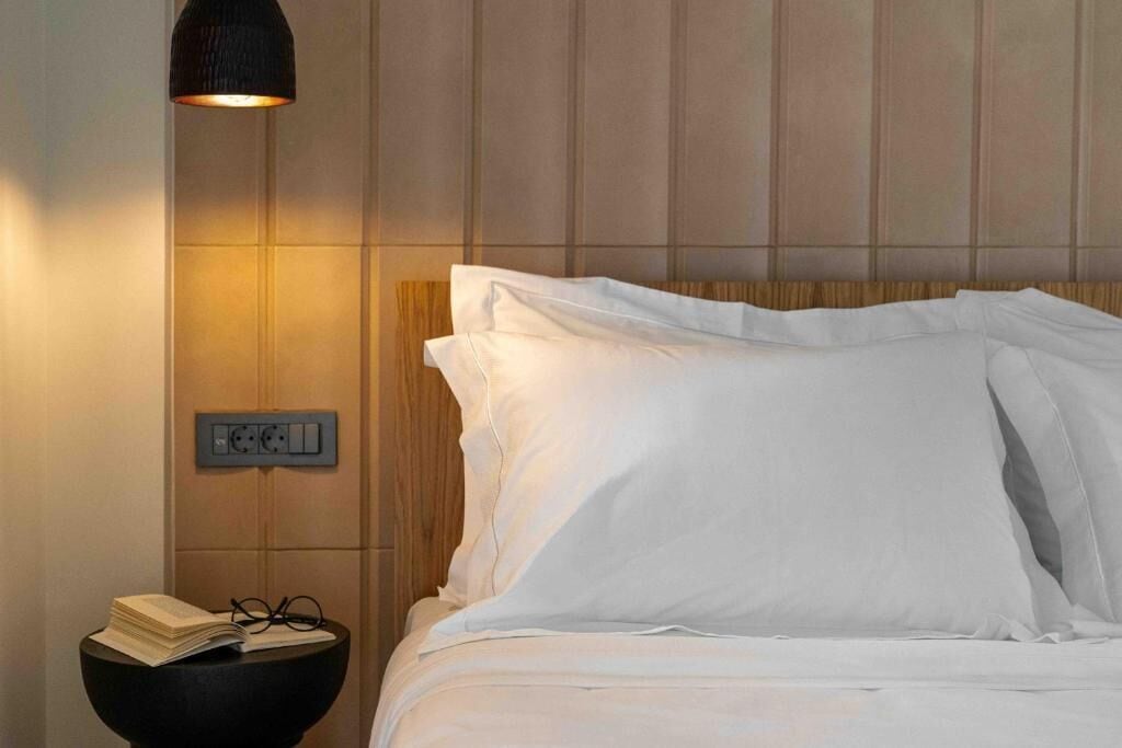 Una cama bien hecha con sábanas blancas, una mesita de noche con un libro y gafas, y un enchufe de pared.