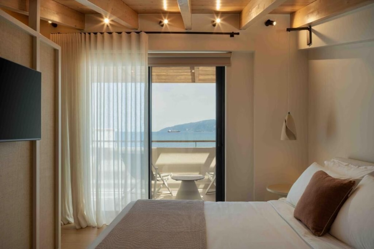 Quarto de hotel com cama, varanda com mesa e cadeiras, e vista para o mar.