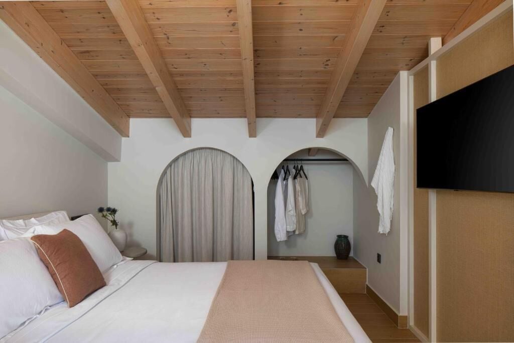A bedroom with a wooden ceiling, a bed with white bedding, a flat-screen TV, and a clothing rack.