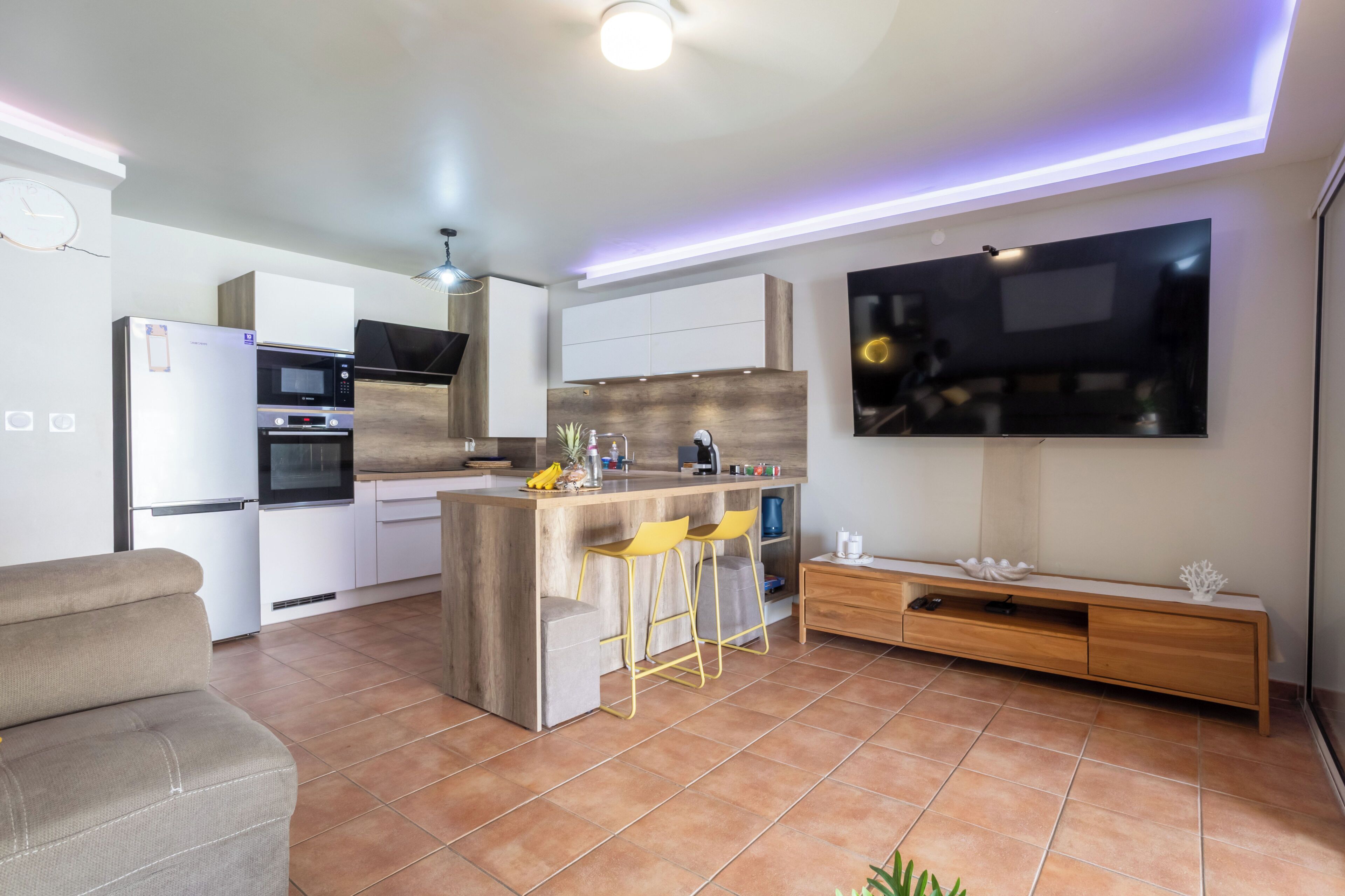 A modern kitchen with a central island, bar stools, a built-in oven, and a flat-screen TV mounted on the wall.