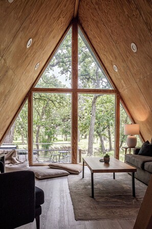A cozy interior with a large window, a sofa, a coffee table, and a lamp. - A-Frame Ranch: Comal Cabin with small pool (Fredericksburg)