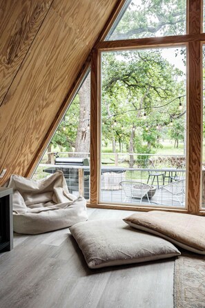 A room with a sloped ceiling, a large window, and a view of trees and a patio area. - A-Frame Ranch: Comal Cabin with small pool (Fredericksburg)