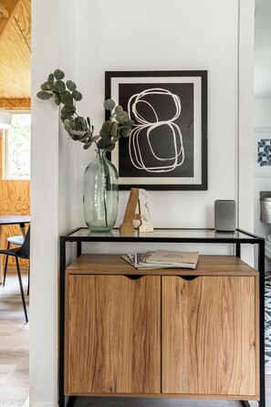 A room with a wooden cabinet, a vase with green leaves, a framed abstract artwork, and a small table with a magazine. - A-Frame Ranch: Comal Cabin with small pool (Fredericksburg)