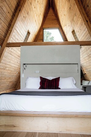 A bedroom with a wooden ceiling and a skylight, a bed with white bedding and a red pillow, and a bedside table with a lamp. - A-Frame Ranch: Post Oak Treehouse with Shared Pool (Fredericksburg)