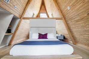 A bedroom with a sloped ceiling, a bed with white bedding, a red pillow, and a bedside table with a lamp. - A-Frame Ranch: Post Oak Treehouse with Shared Pool (Fredericksburg)
