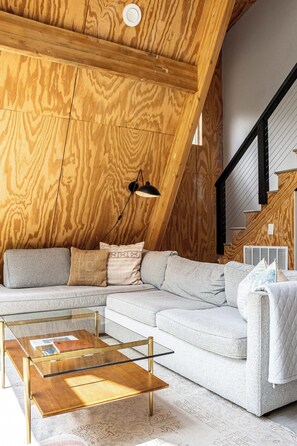 A modern loft-style living space with a wooden sloped ceiling, a sectional sofa, a glass coffee table, and a staircase with a metal railing. - A-Frame Ranch: Llano Cabin with Cowboy Pool (Fredericksburg)