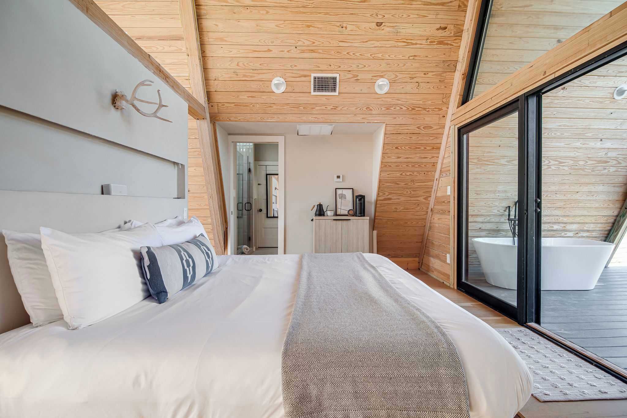A modern bedroom with a large bed, a wooden ceiling, and a sliding glass door leading to a bathroom with a bathtub.
