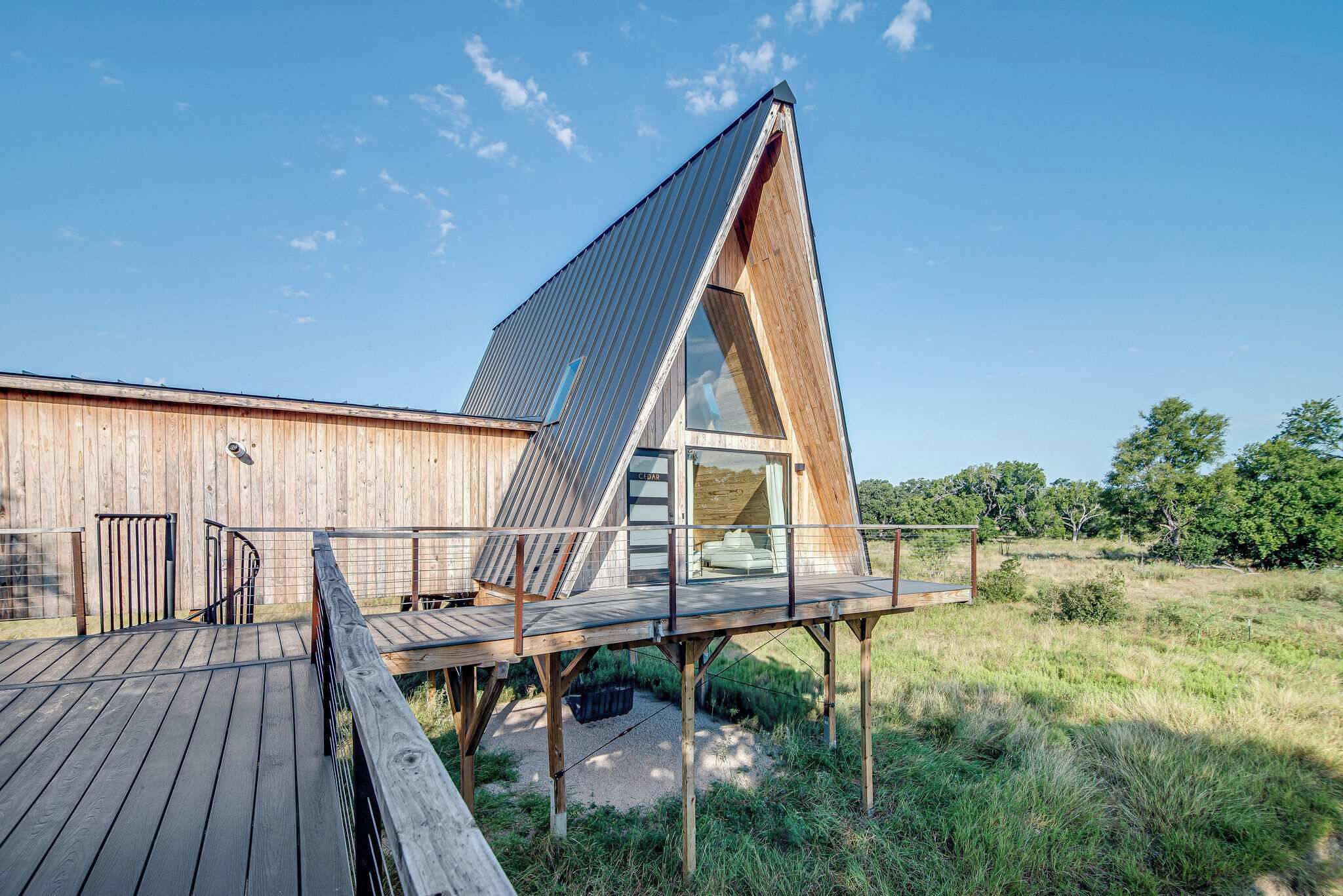 A modern, triangular wooden structure with a glass balcony, elevated on stilts, adjacent to a wooden building.