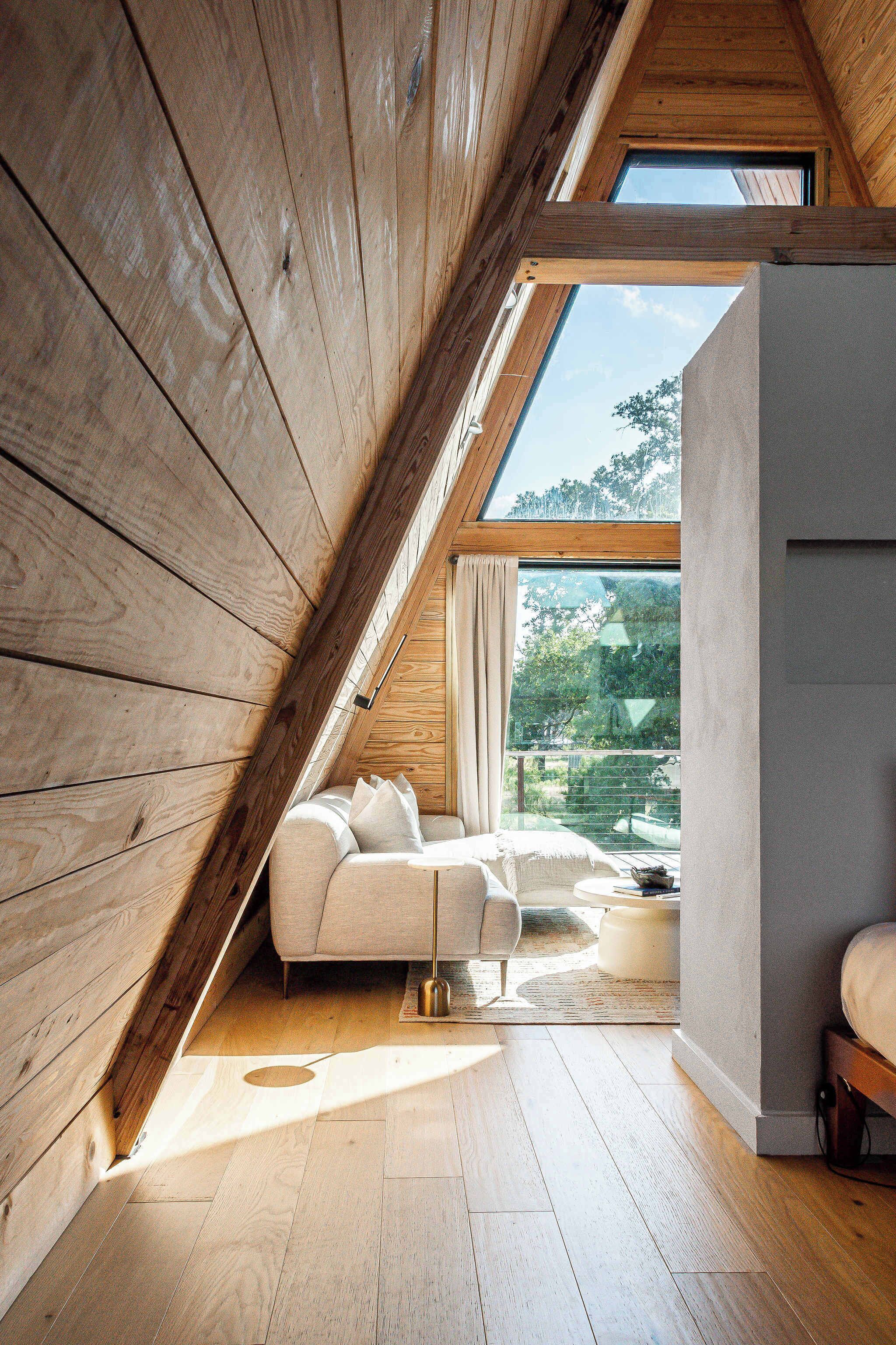 A modern loft bedroom with a sloped ceiling, wooden beams, a large window, and a sofa.